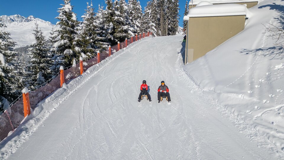 Zwei Personen sitzen auf einer verschneiten Piste. Umgeben von schneebedeckten Bäumen und einem klaren blauen Himmel.
