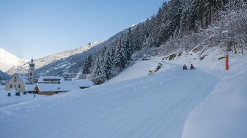 Eine winterliche Landschaft mit Schnee bedeckten Hügeln und Tannenbäumen. Im Hintergrund sind kleine Häuser und eine Kirche zu sehen.