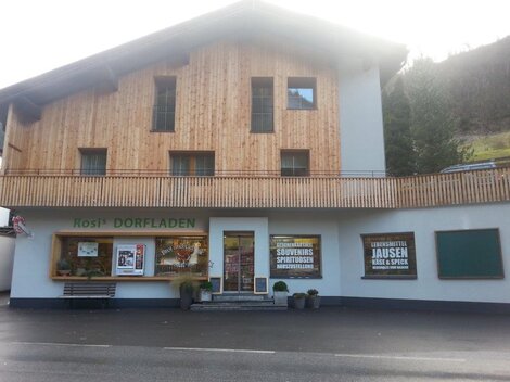 Ein traditioneller Dorfladen mit Holzfassade und einer einladenden Eingangsfront. Vor dem Laden befinden sich Werbeplakate für lokale Produkte und Spezialitäten.