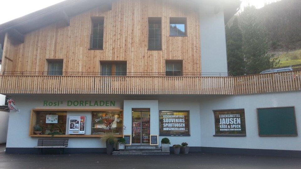 Ein traditioneller Dorfladen mit Holzfassade und einer einladenden Eingangsfront. Vor dem Laden befinden sich Werbeplakate für lokale Produkte und Spezialitäten.
