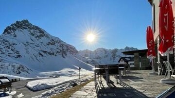 Eine sonnige Terrasse mit Blick auf schneebedeckte Berge. Im Vordergrund stehen Stühle und Tische, die zum Entspannen einladen.