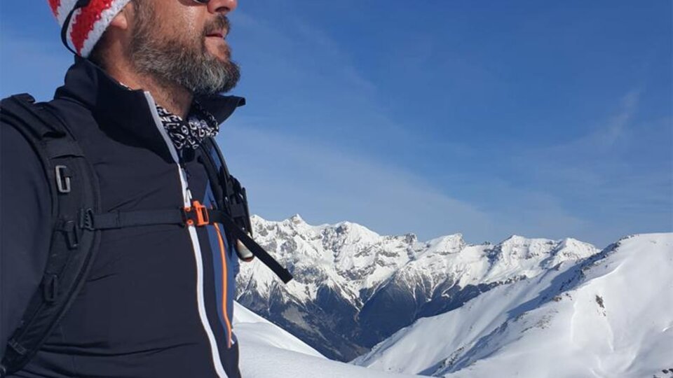 Ein Mann mit Sonnenbrille und Strickmütze steht auf einem schneebedeckten Berggipfel. Im Hintergrund sind majestätische, schneebedeckte Berge unter einem klaren blauen Himmel zu sehen.