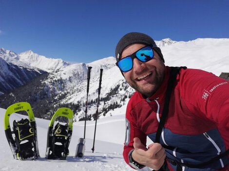 Ein Mann macht ein Selfie im Schneegebirge. Im Hintergrund sind verschneite Berge und Schneeschuhe zu sehen.
