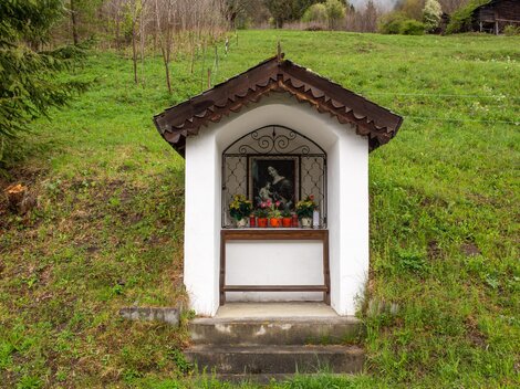 Eine kleine Kapelle auf einem grünen Hügel. Vor der Kapelle steht ein Fenster mit Blumen und einem Bild.