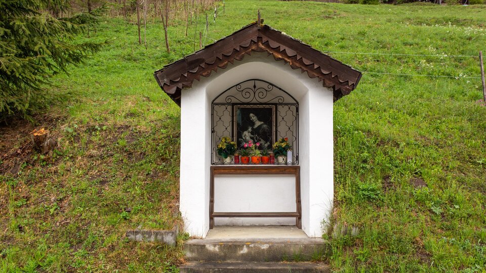 Eine kleine Kapelle auf einem grünen Hügel. Vor der Kapelle steht ein Fenster mit Blumen und einem Bild.