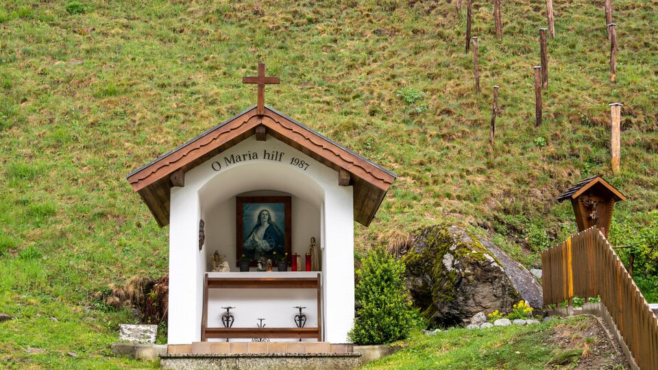Eine kleine Kapelle mit einem Kreuz und einem Bild der Jungfrau Maria. Sie steht inmitten einer grünen Wiese.