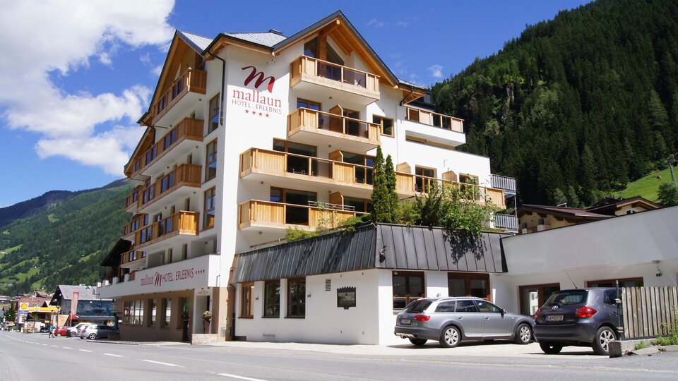 Ein modernes Hotelgebäude mit mehreren Etagen und Balkonen. Umgeben von grünen Bergen und klarem Himmel.