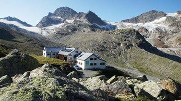Eine wunderschöne Berglandschaft mit schroffen Felsen und hohen Gipfeln. Im Vordergrund stehen zwei Hütten, eingebettet in die Natur.